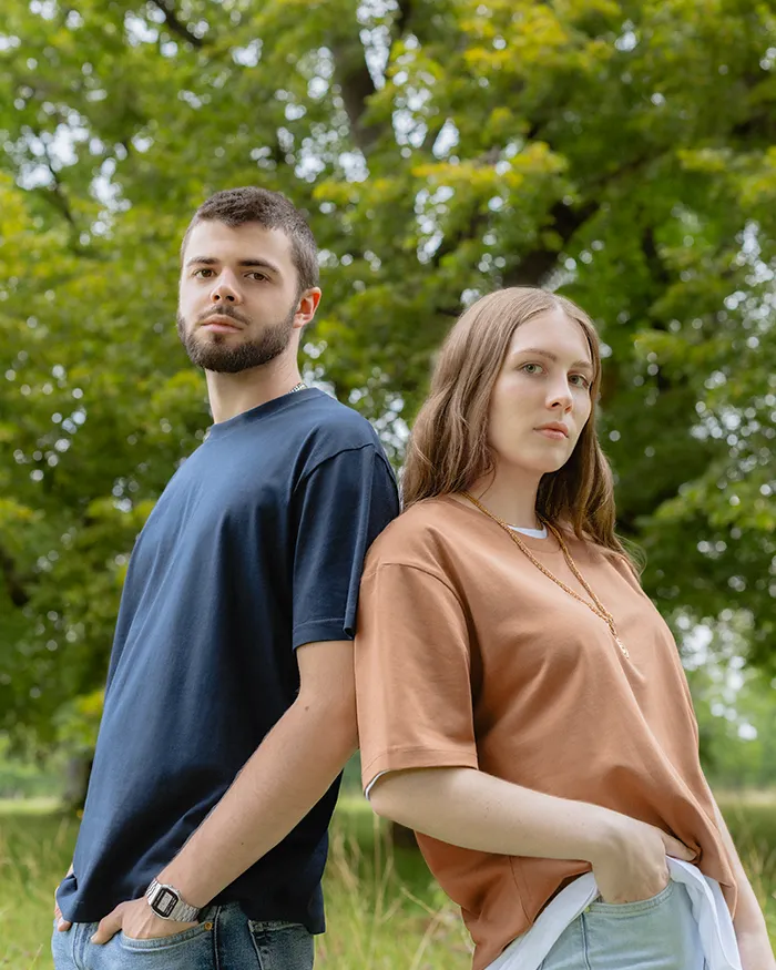Ein Mann mit dunklem Haar und Bart in einem blauen T-Shirt und eine blonde Frau in einem braunen T-Shirt die aneinander lehnen vor einem großen Baum