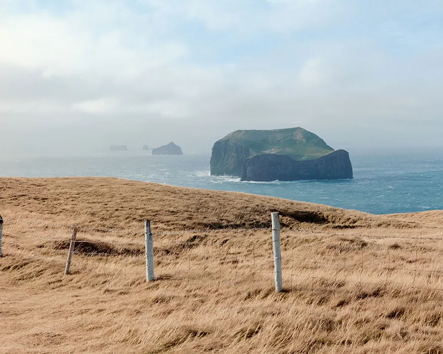 Aussicht von der Westmännerinseln mit einer Wiese und Zaun im Vordergrund und kleineren Inseln auf dem Meer